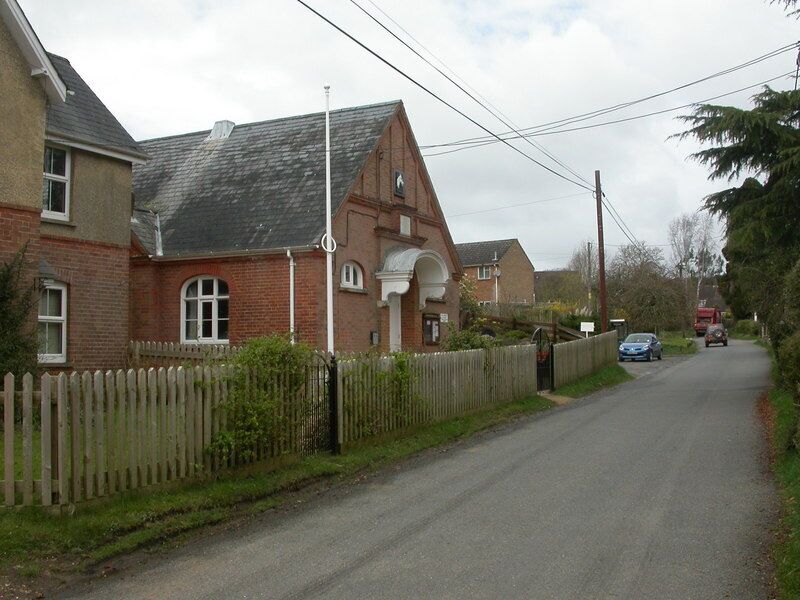 A narrow residential street with houses on both sides, a wooden fence, and a car parked on the right.