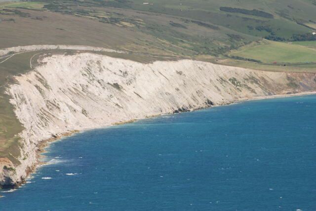 A coastal cliff with a steep drop into the ocean, surrounded by green hills.