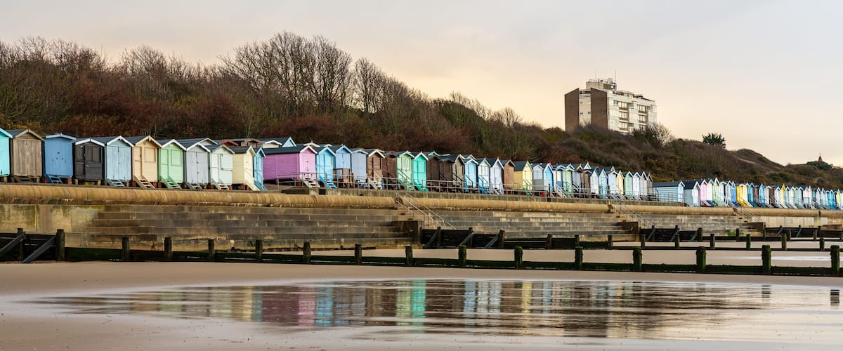 A row of colorful beach huts along a waterfront, with a building on a hill in the background.