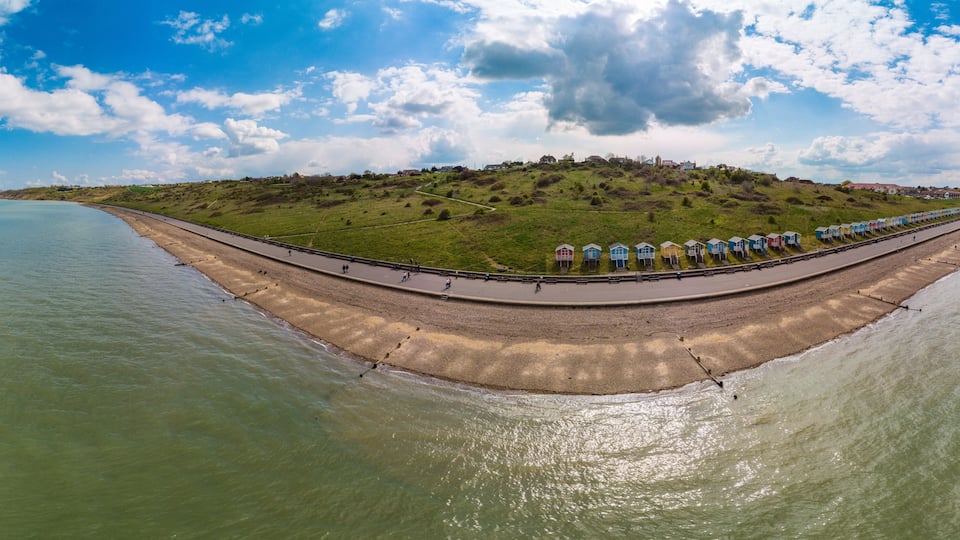 A coastal landscape with a curved promenade, beach houses, and a grassy hill in the background.