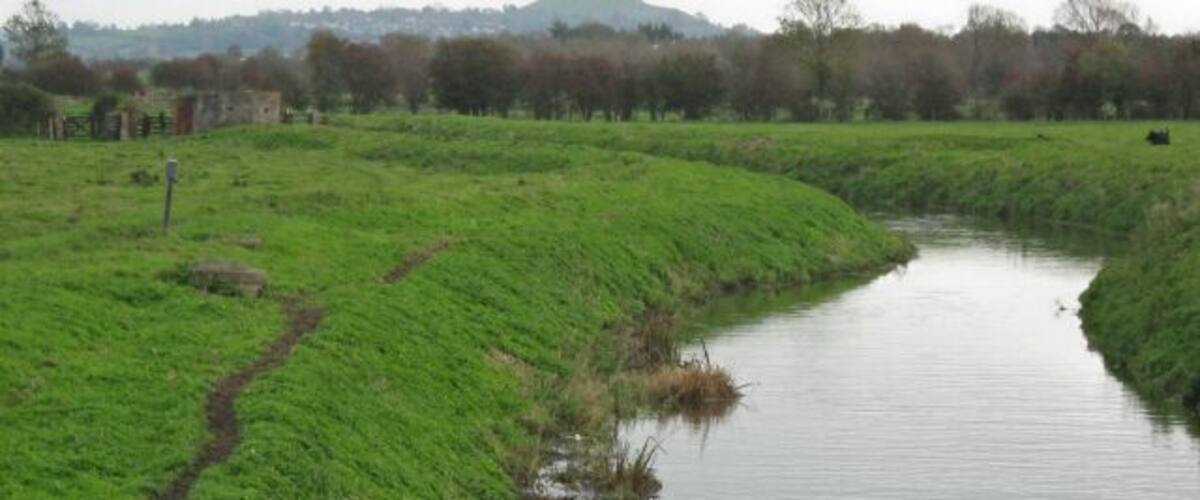 A narrow water channel with grassy banks, a path, and a distant hill.