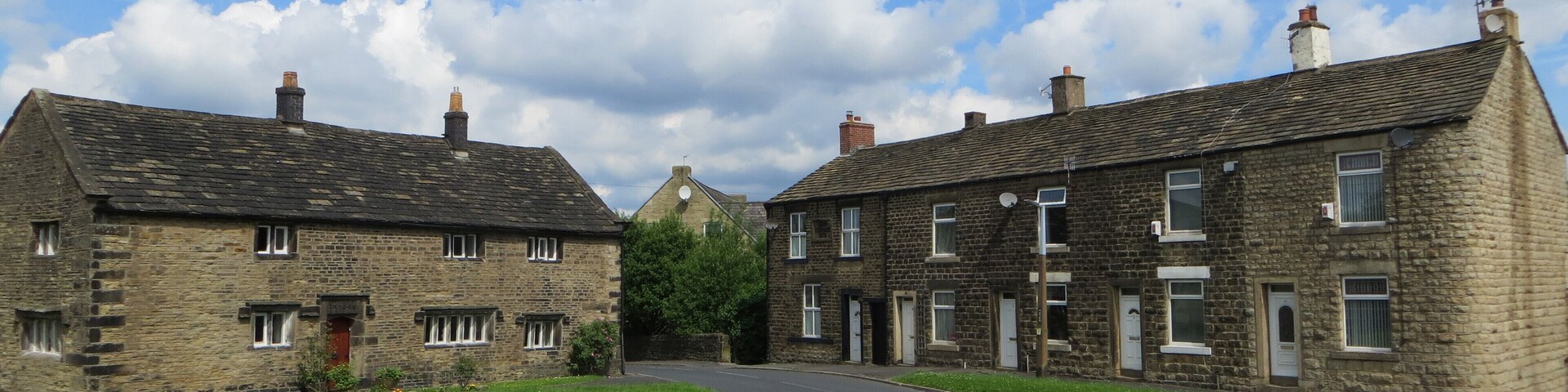 A rural road with stone buildings on either side, surrounded by green fields and a partly cloudy sky.