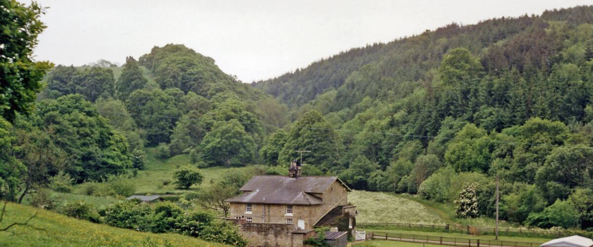 A stone house with a watermill in a lush green valley surrounded by forested hills.