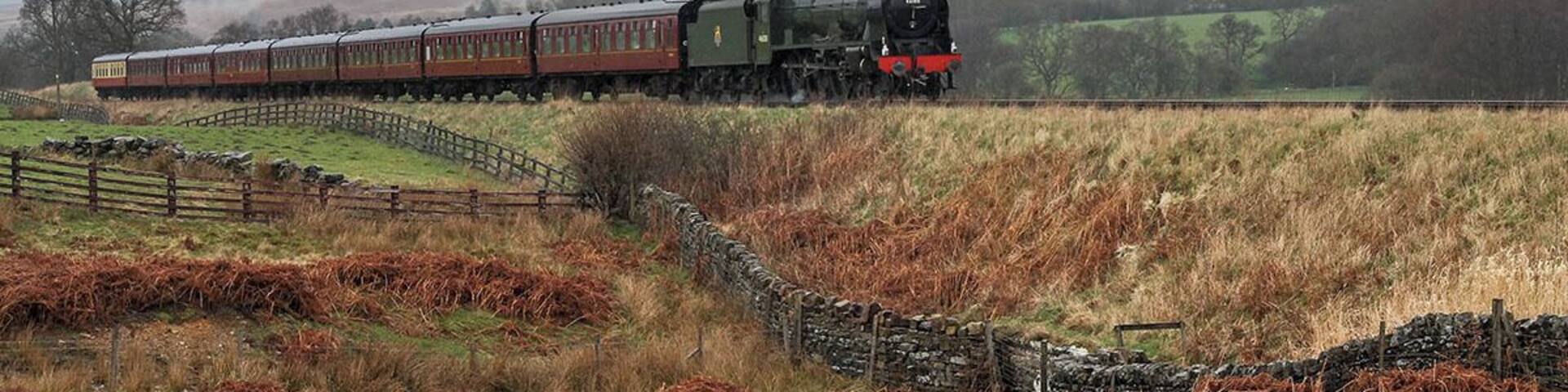 A steam locomotive with a large plume of steam, traveling through a rural landscape with stone walls and grassy fields.