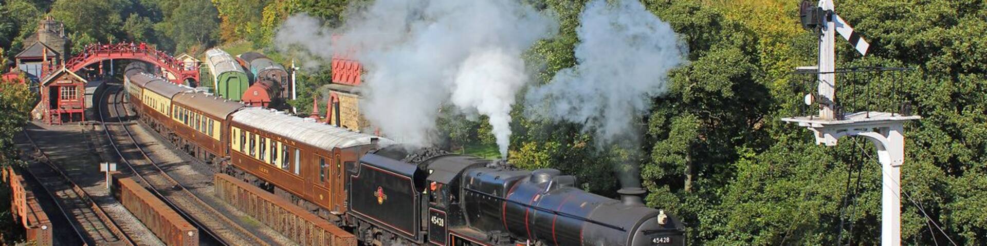 A steam locomotive on a railway track surrounded by lush greenery and a red bridge.