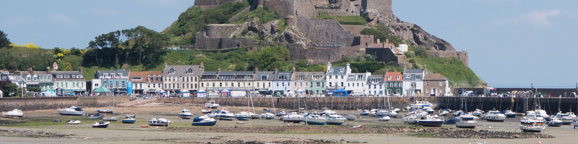 Un pueblo costero con un castillo imponente en una colina, una playa de arena y numerosos barcos amarrados a la orilla.