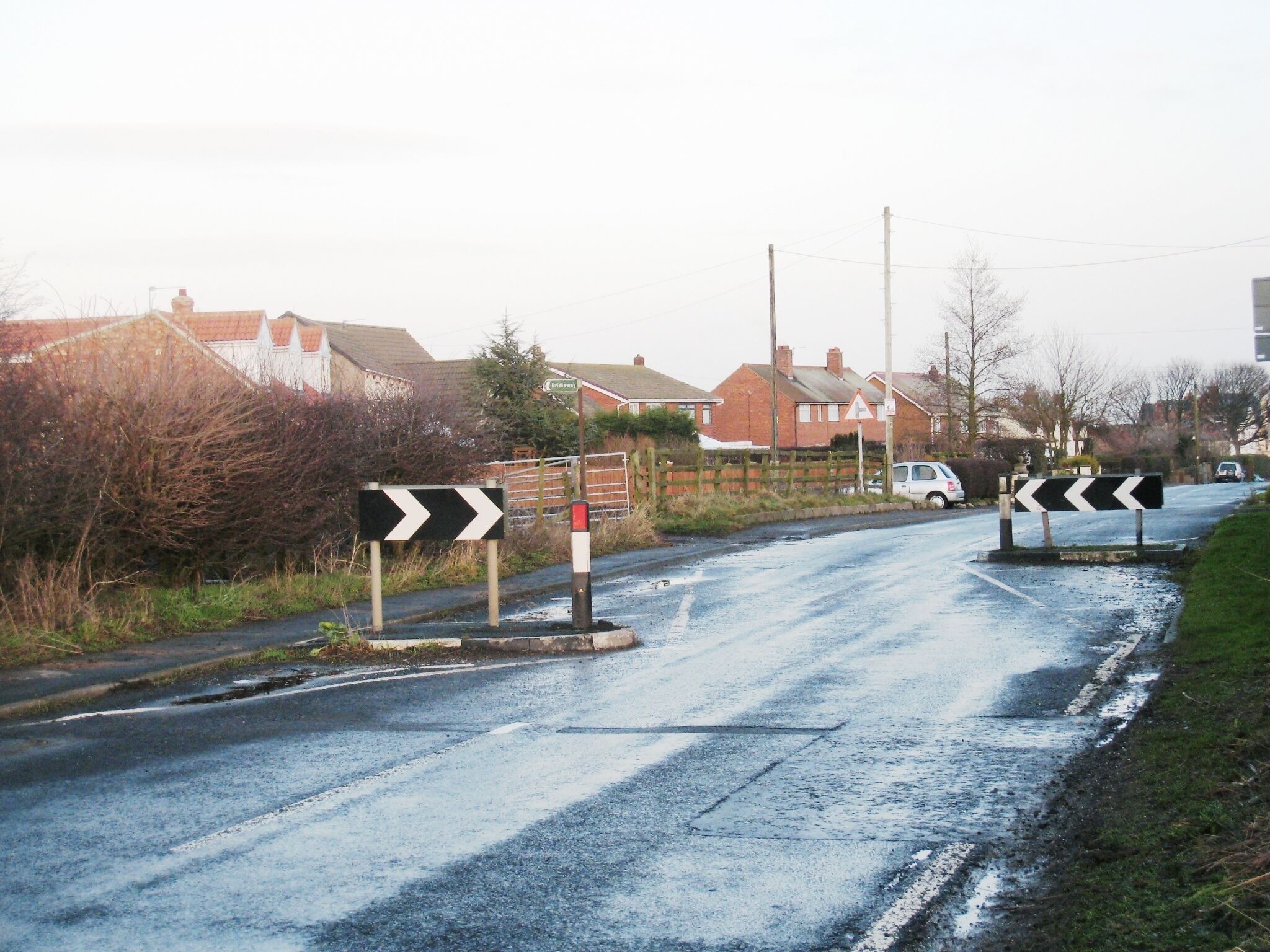 A rural road with chevron signs indicating a sharp bend to the right. Houses line the road, and a car is parked on the side.