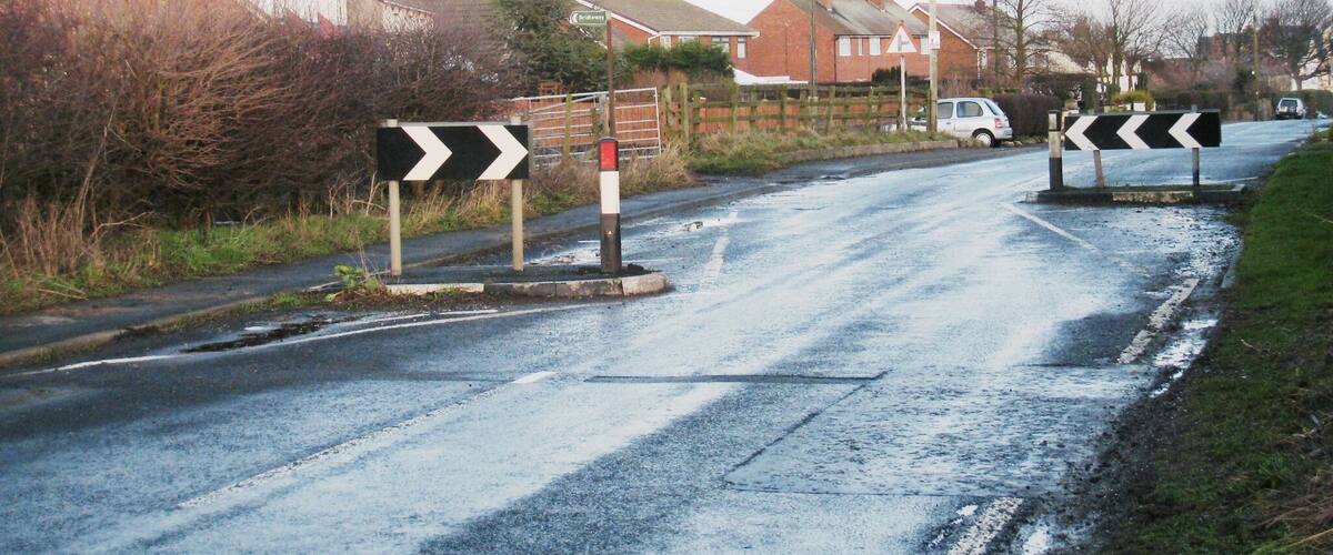 A rural road with chevron signs indicating a sharp bend to the right. Houses line the road, and a car is parked on the side.