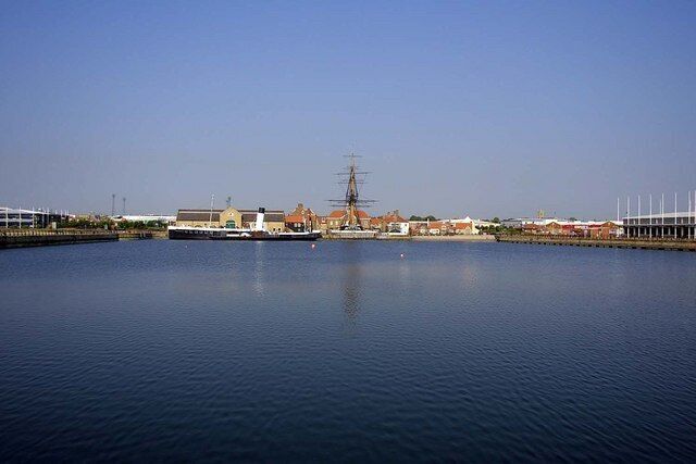 A calm harbor with a large ship, buildings, and a clear sky.
