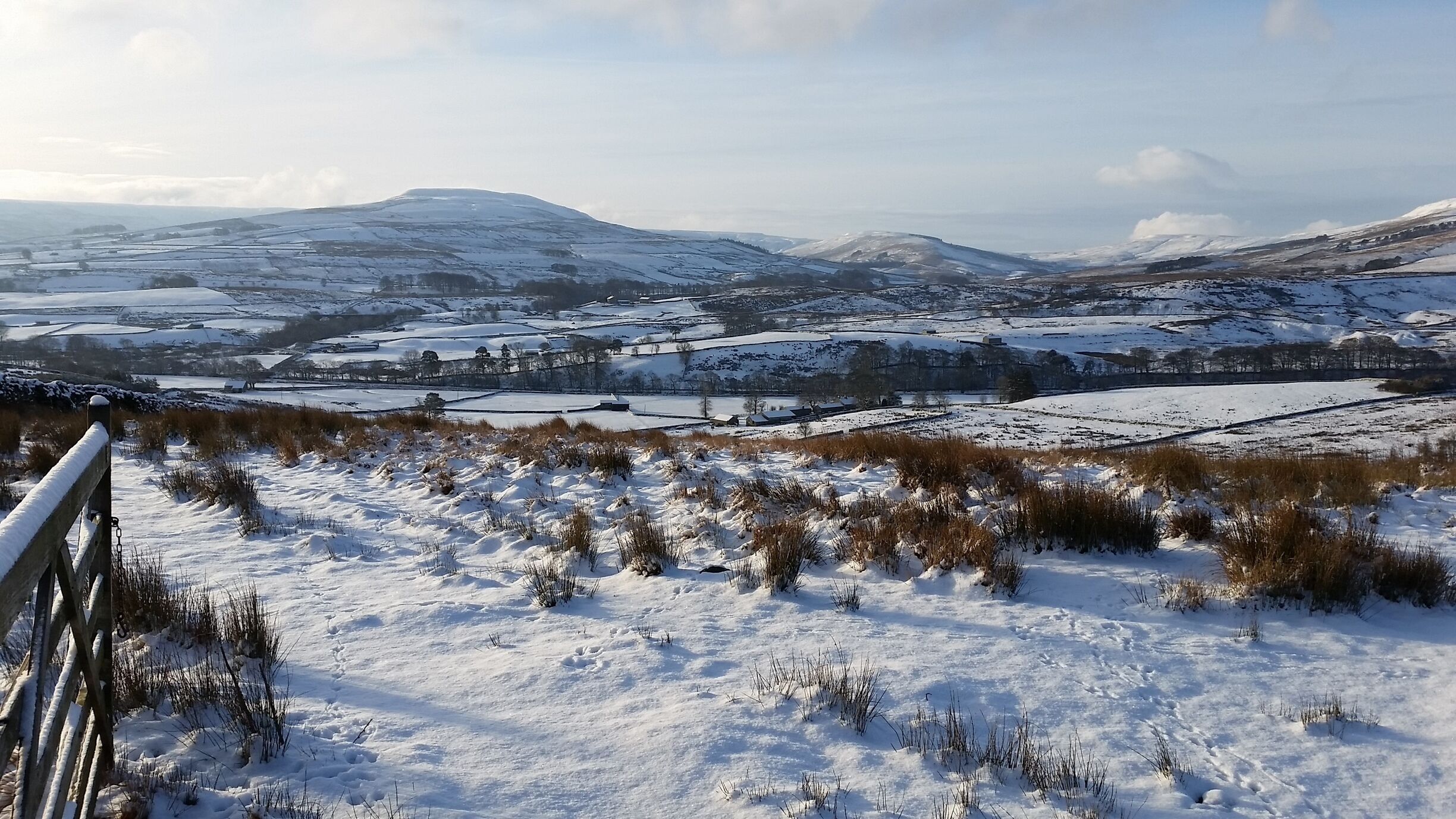 A snow-covered landscape with fields, a fence, and distant mountains.
