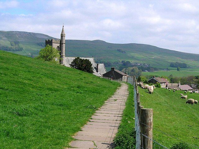 A grassy path leading to a stone church with a tower, surrounded by sheep and rolling hills.