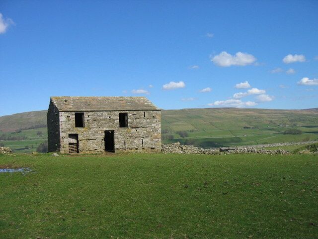 A stone building with two doors and windows, surrounded by a grassy field and a stone wall, with hills in the background.