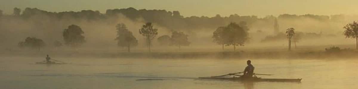 Ein ruhiger See mit einer in der Ferne rudelnden Person, umgeben von Nebel und Bäumen.