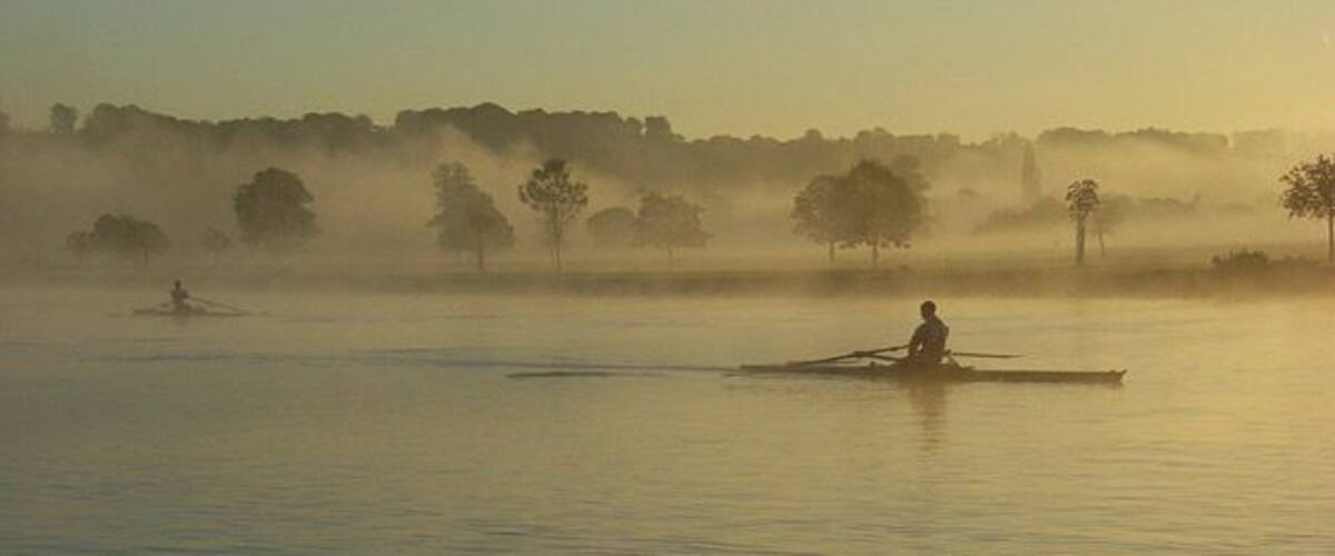 A calm lake with a person rowing a boat in the distance, surrounded by mist and trees.