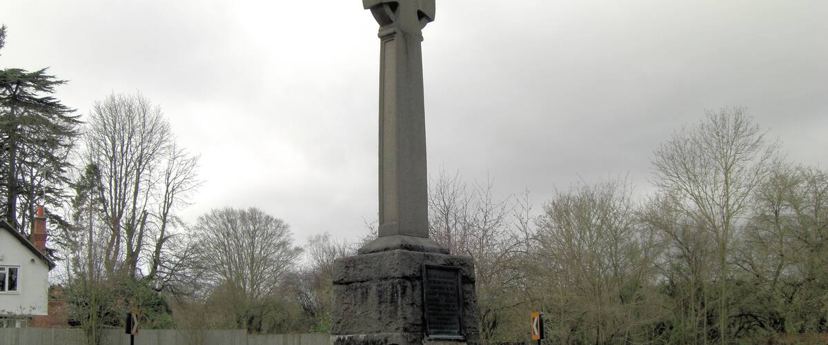 A tall stone monument in a grassy field with trees and a house in the background.