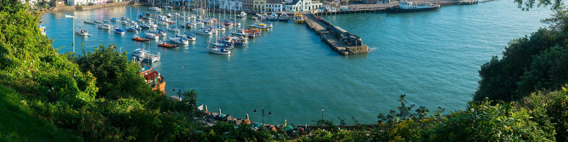 A coastal town with a harbor filled with boats, a lighthouse, and a hill with a fort on top.