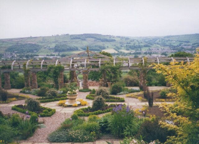 A garden with a fountain, a wooden bridge, and rolling hills in the background.