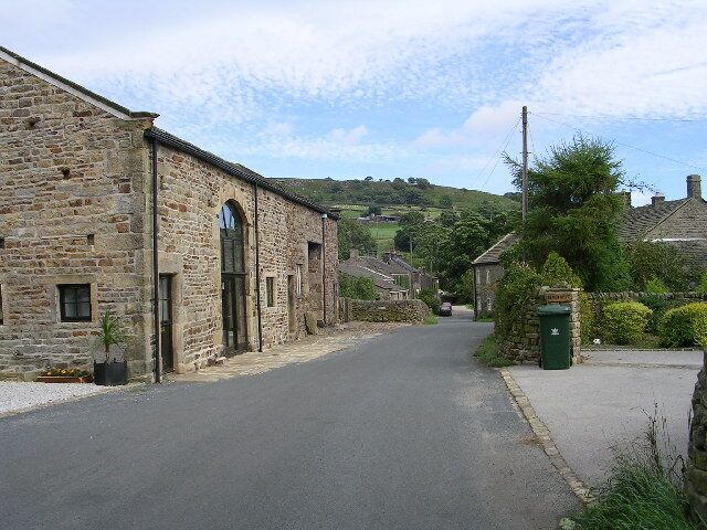 A narrow stone road in a rural village with stone buildings and a green hill in the background.