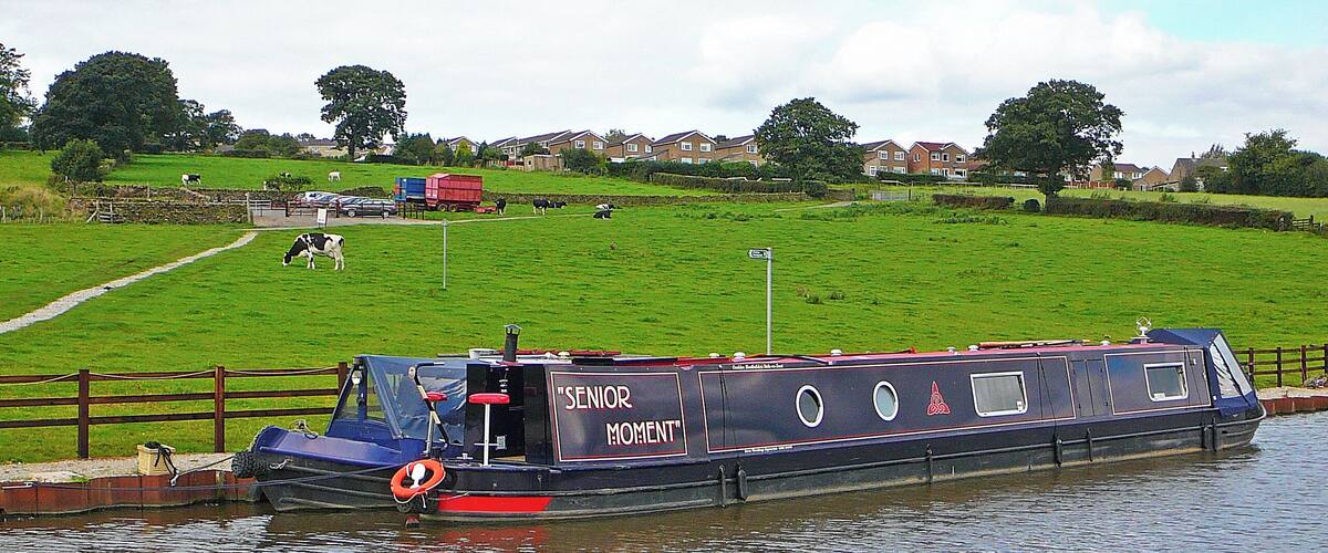 A narrowboat named "SENIOR MOMENT" docked at a canal with a grassy field and houses in the background.