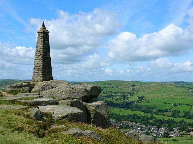 A stone obelisk on a grassy hill with a village in the valley below.