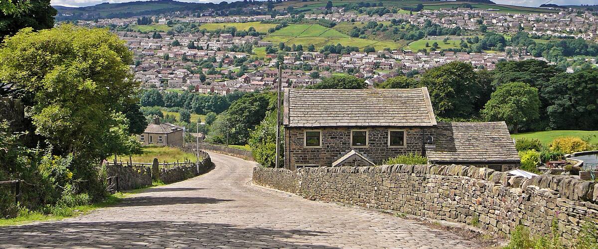 A cobblestone path leading to a stone building with a thatched roof, surrounded by lush greenery and a view of a town in the distance under a cloudy sky.