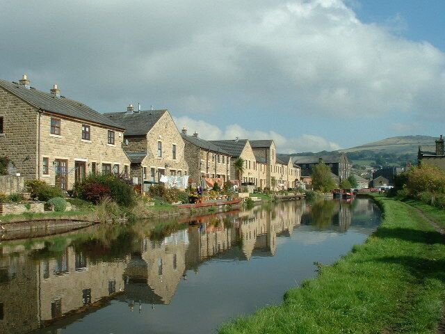 A river reflecting stone buildings, a grassy bank, and a bridge.