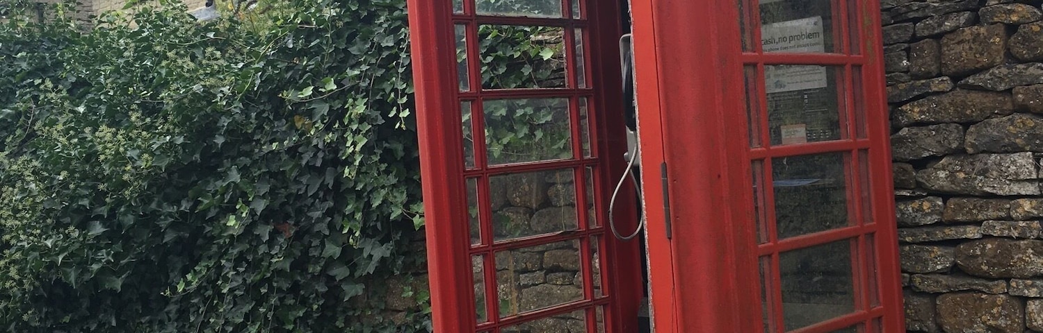 A red British telephone booth with a stone wall and greenery in the background.
