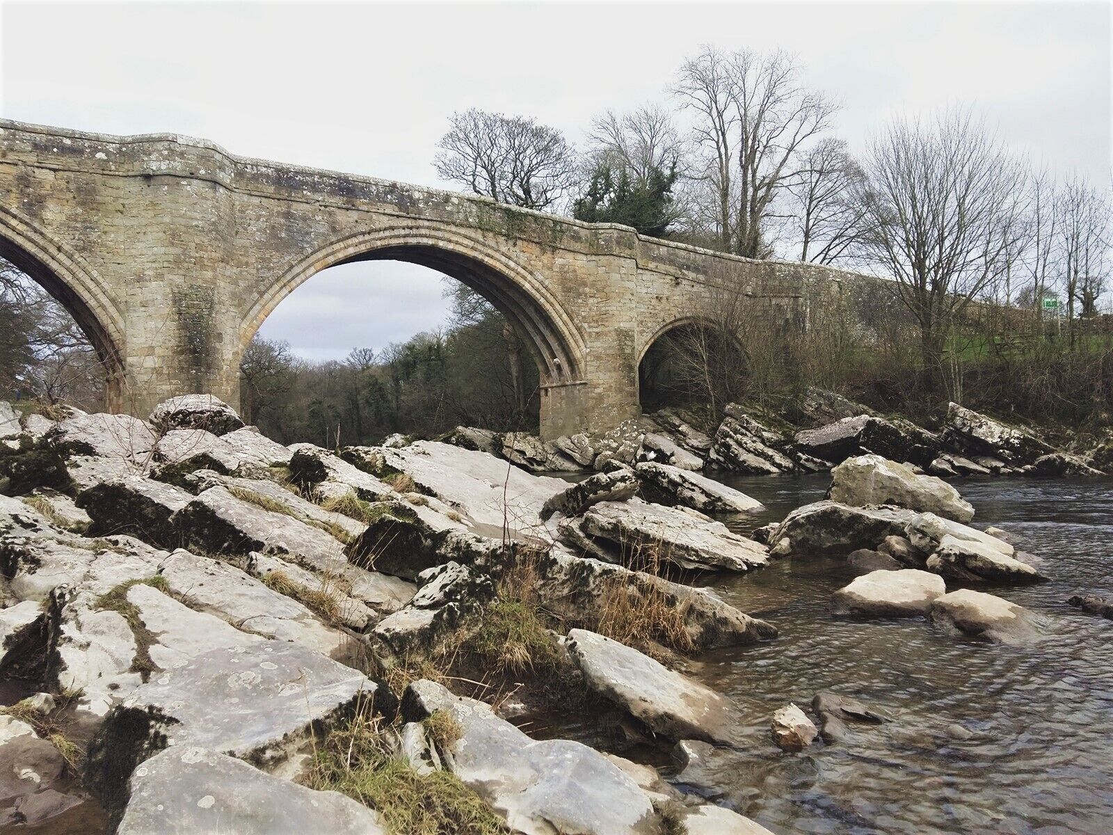 A stone bridge over a rocky riverbed with trees in the background.