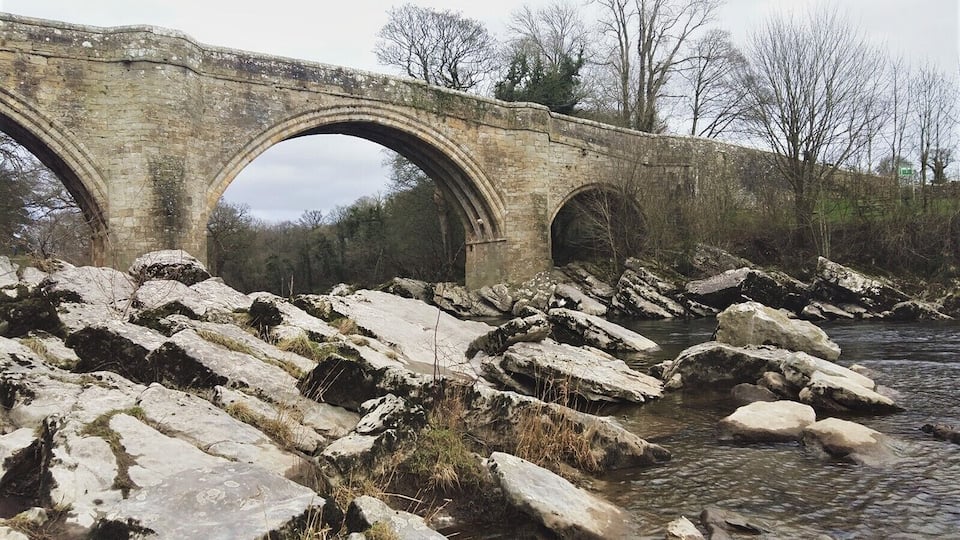 A stone bridge over a rocky riverbed with trees in the background.