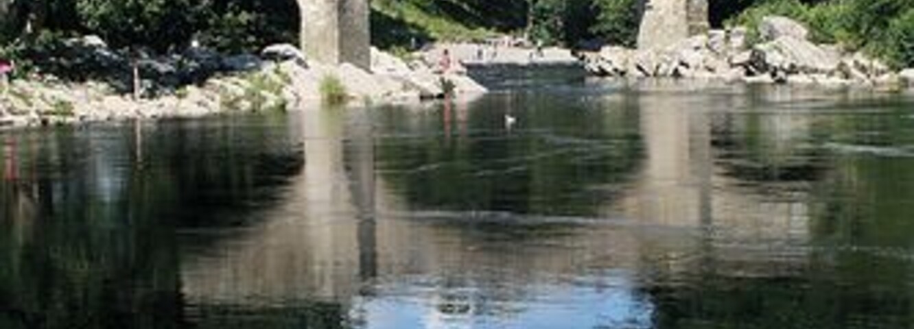 Un puente de piedra sobre un río tranquilo con reflejos nítidos, rodeado de árboles y vegetación.