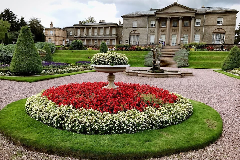 A formal garden with a central circular flower bed, a fountain, and a classical building in the background.