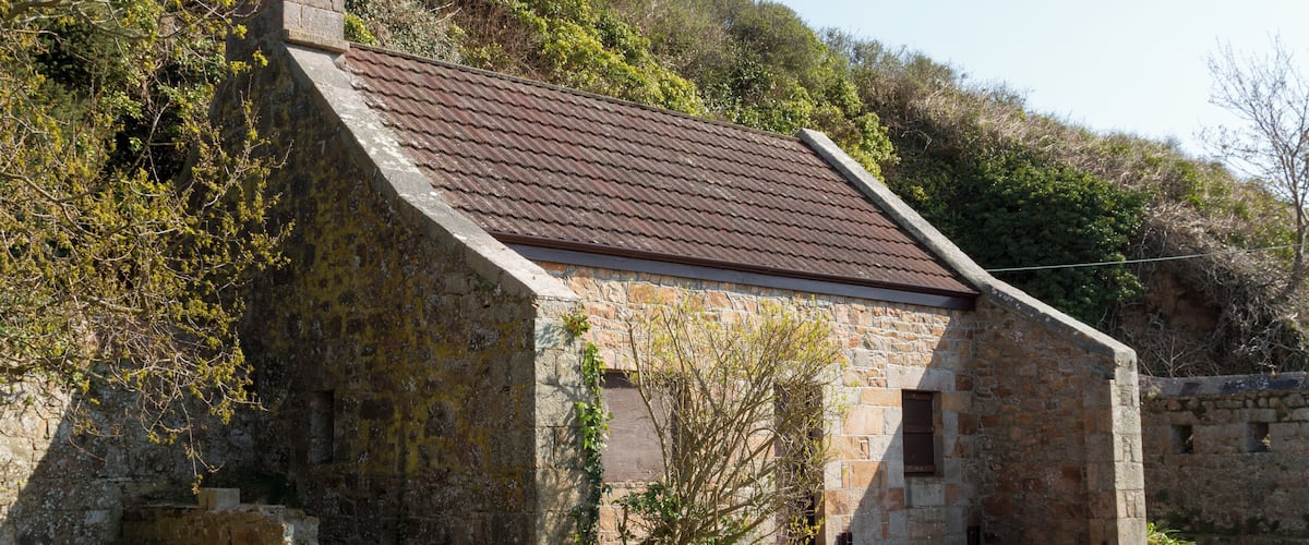A stone building with a sloped roof, surrounded by greenery and hills.