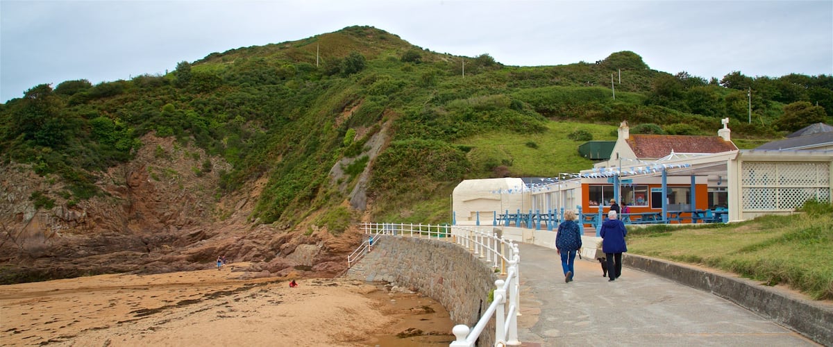 A coastal walkway with a sandy beach, a stone bridge, and a hill with greenery.