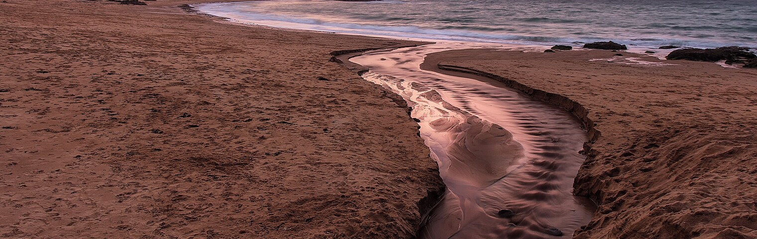 Uma praia com um riacho sinuoso, um penhasco rochoso e um céu ao pôr do sol.