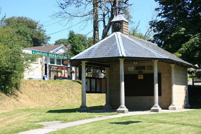 A gazebo with a clock, surrounded by grass and trees, near a shop.