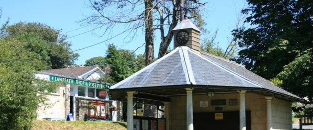 A gazebo with a clock, surrounded by grass and trees, near a shop.