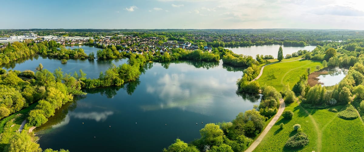 A river winding through a park with a path, surrounded by lush greenery and a town in the background.