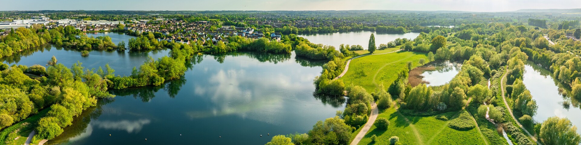 A river winding through a park with a path, surrounded by lush greenery and a town in the background.