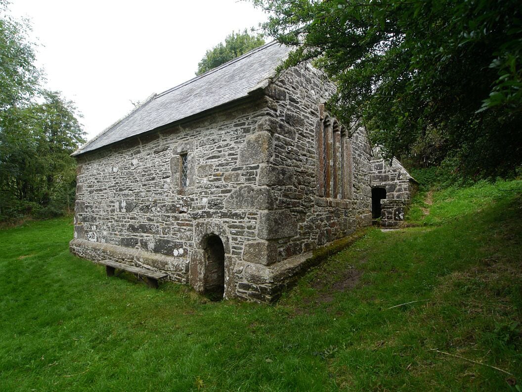 A stone church with a sloped roof, surrounded by lush greenery.