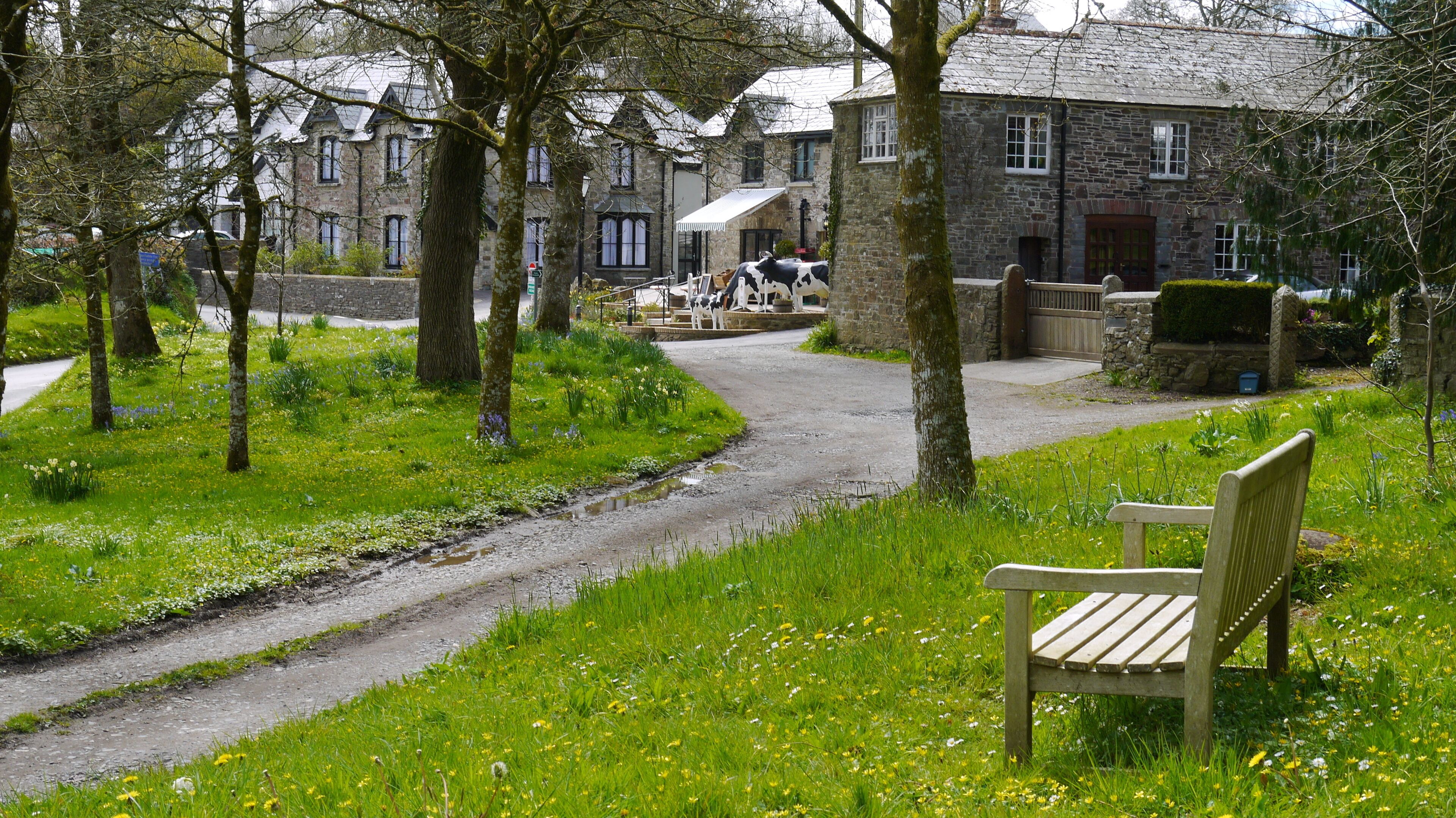A rural pathway with a bench, lush greenery, and stone buildings in the background.