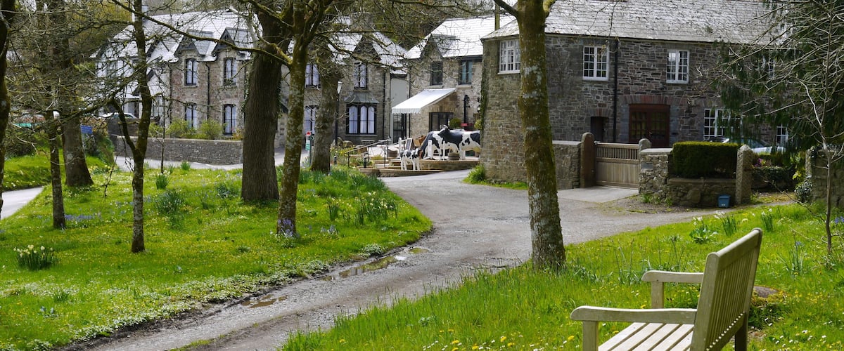 A rural pathway with a bench, lush greenery, and stone buildings in the background.