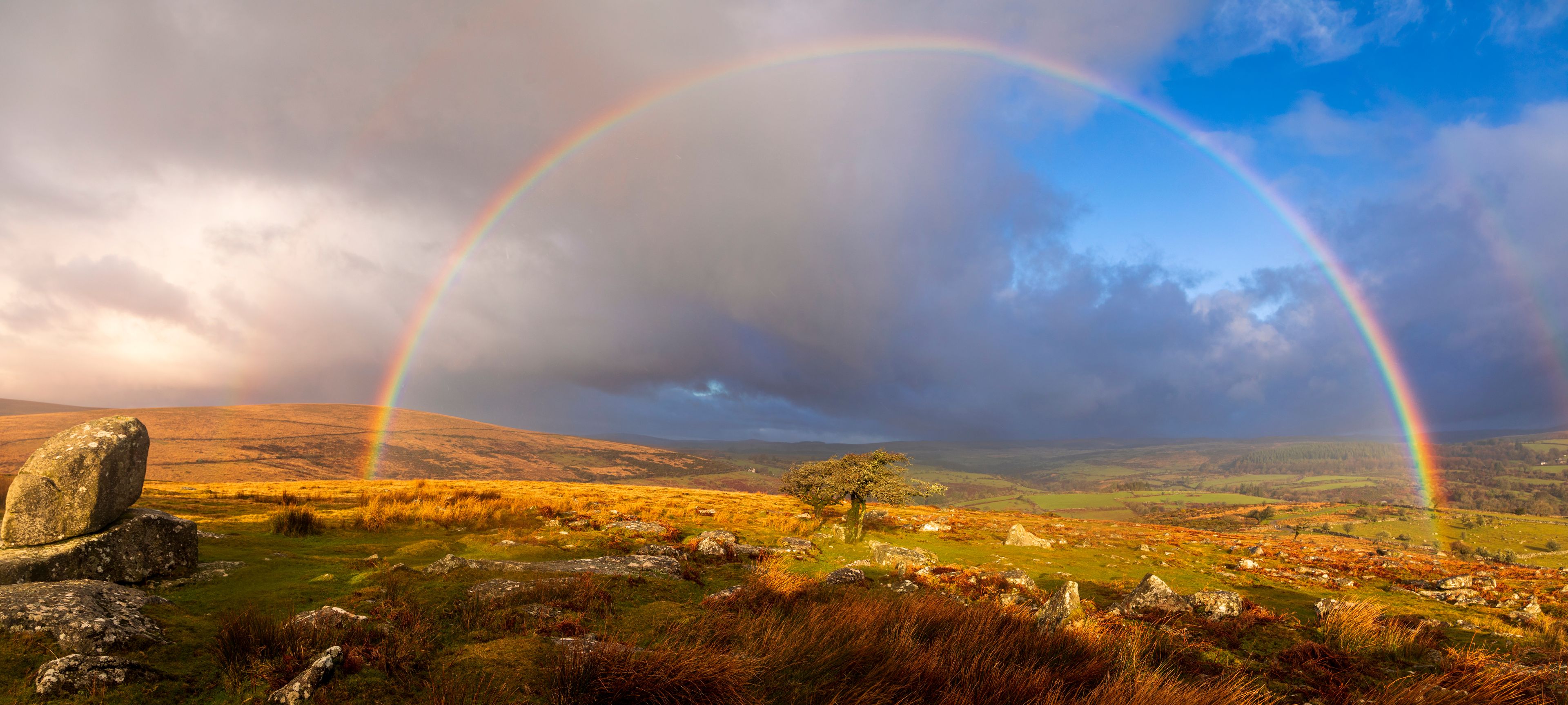 Un paysage avec deux arcs-en-ciel, un cercle de pierres et un arbre isolé.