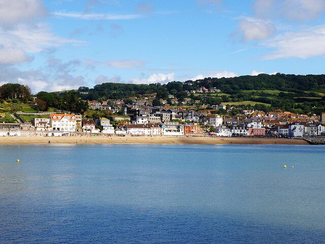 A coastal town with buildings along the shore, a beach, and a hill with greenery in the background.
