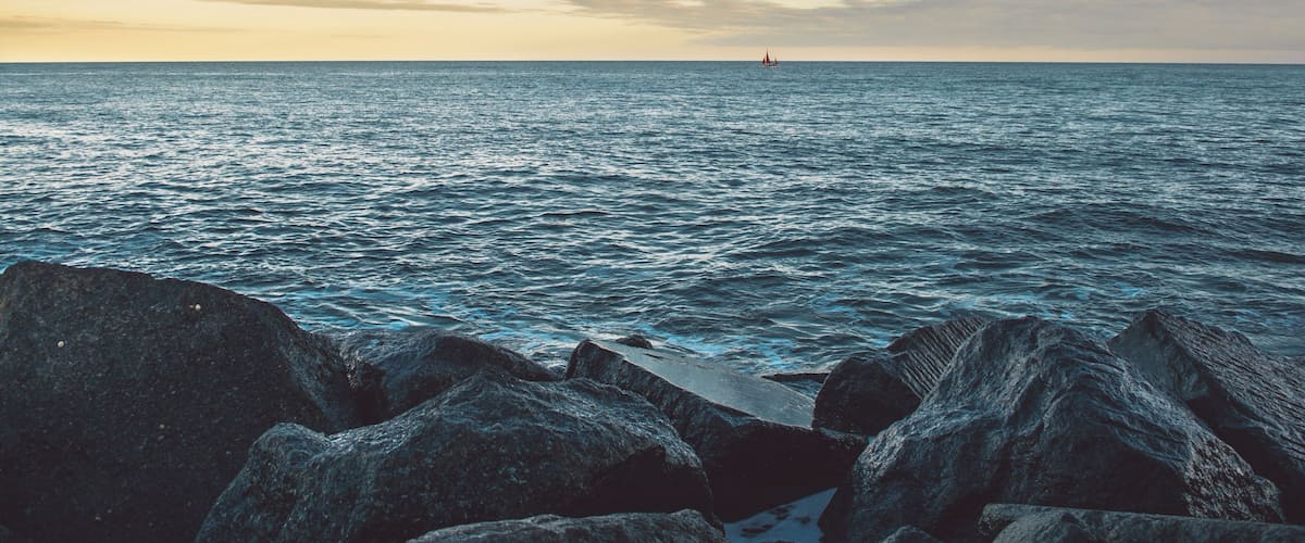 A rocky shoreline with calm sea and a distant sailboat.