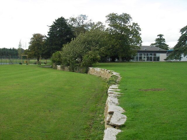 Un campo cubierto de césped con un muro de piedra,  árboles y un edificio al fondo.