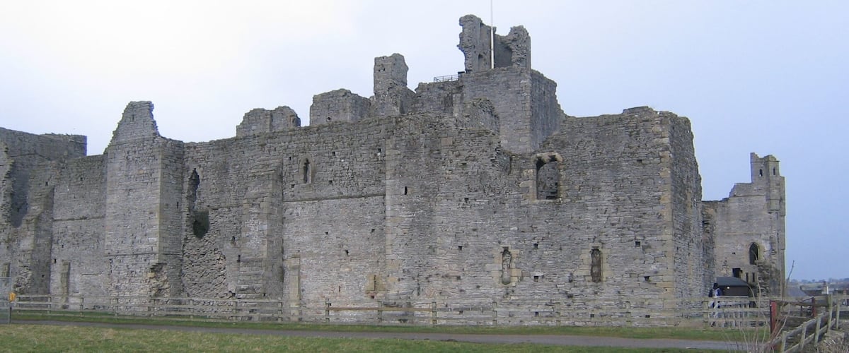 A historic stone castle with multiple towers and battlements, surrounded by a grassy field and a stone wall.
