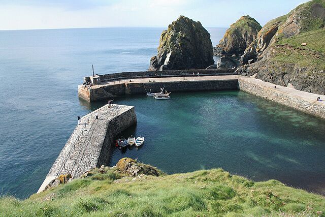 A coastal harbor with a large rock formation, a stone pier, and a small boat anchored near the shore.