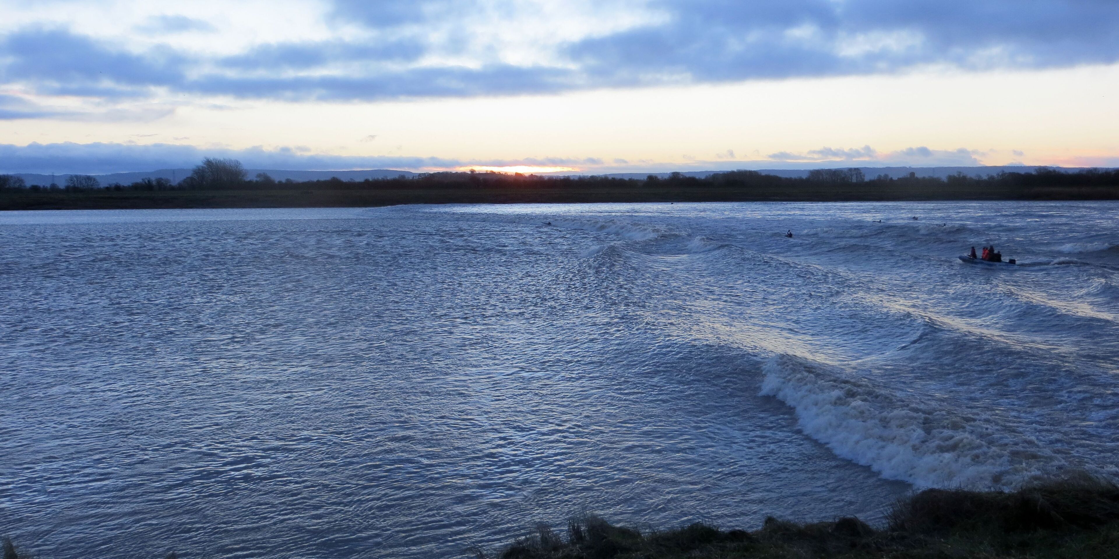 A body of water with waves, a sunset in the background, and a distant shoreline.