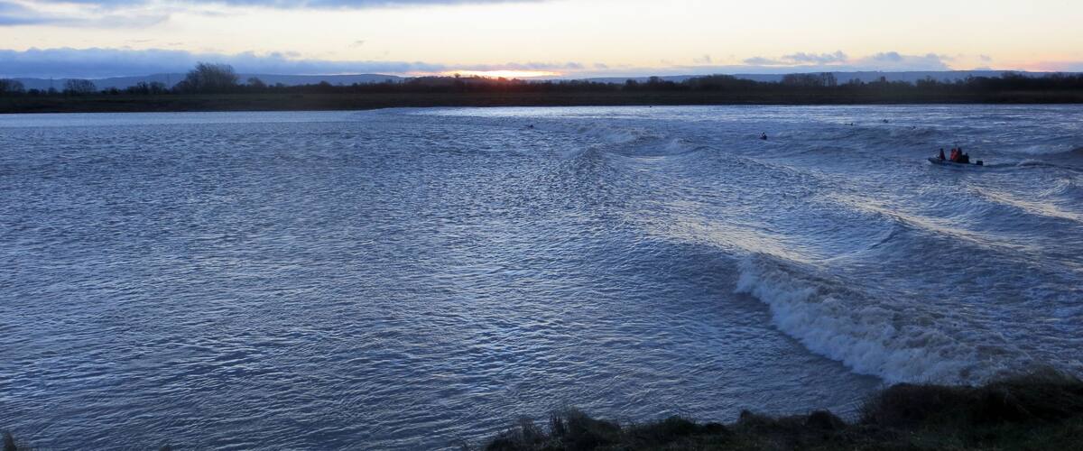 A body of water with waves, a sunset in the background, and a distant shoreline.