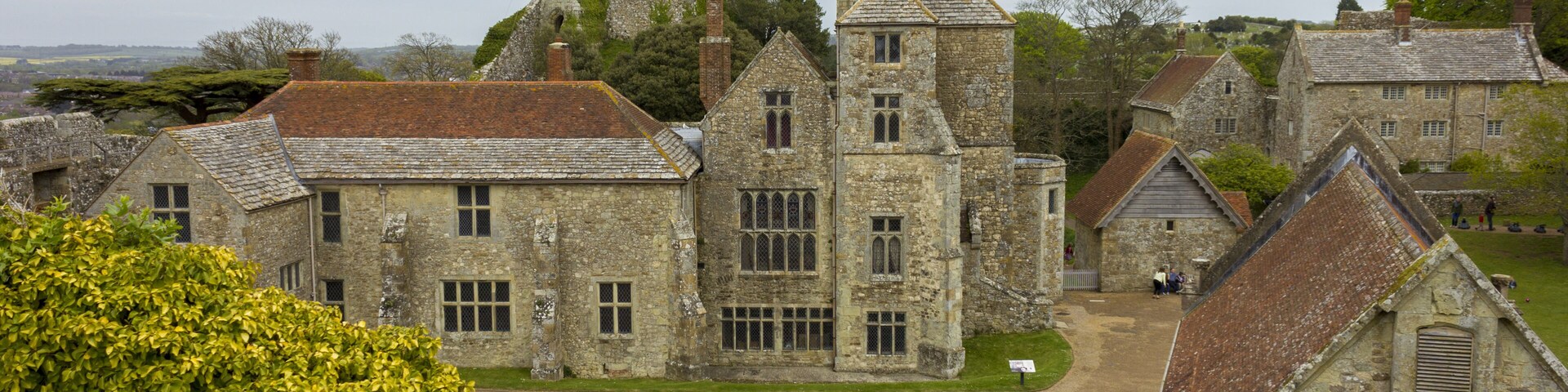 A historic stone building complex with a prominent tower, surrounded by lush greenery and a clear sky.